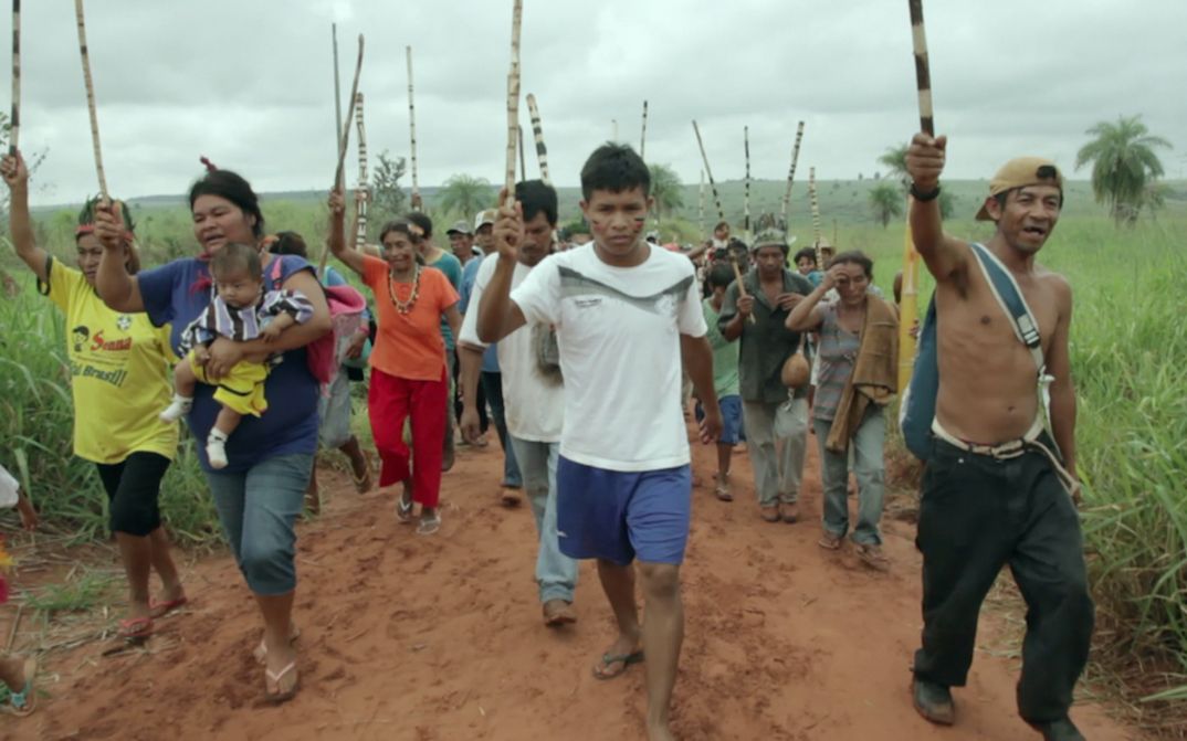 Filmstill aus MARTÍRIO: Eine Gruppe von Menschen geht mit erhobenen Stöcken einen Feldweg entlang.