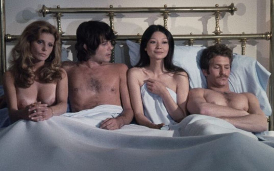 Filmstill aus GROUP MARRIAGE: Zwei Männer und zwei Frauen sitzen und liegen nebeneinander und unbekleidet in einem großen Bett.