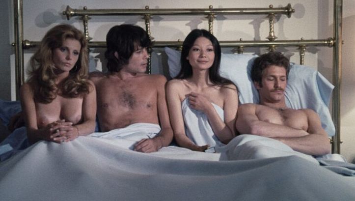 Film still from GROUP MARRIAGE: Two men and two women sit and lie unclothed in a large bed.