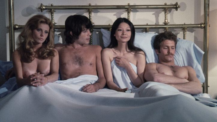 Filmstill aus GROUP MARRIAGE: Zwei Männer und zwei Frauen sitzen und liegen nebeneinander und unbekleidet in einem großen Bett.