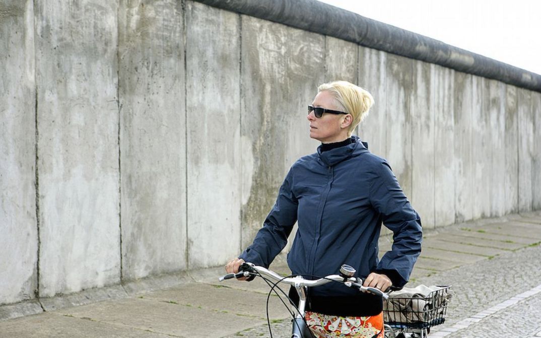 Filmstill aus THE INVISIBLE FRAME: Die Schauspielerin Tilda Swinton steht mit dem Fahrrad vor einem Stück der Berliner Mauer.