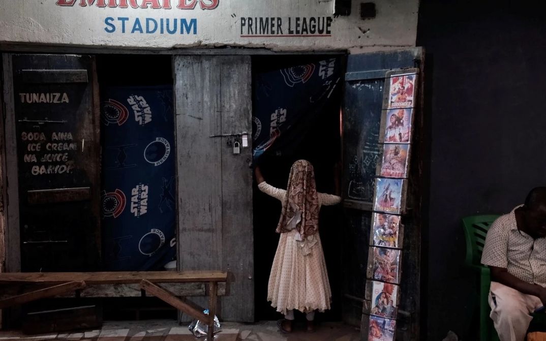 Film still from APOSTLES OF CINEMA: A child enters a simple store with movie posters hanging on the door.
