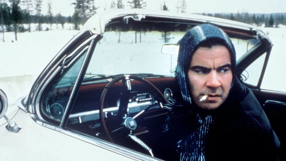 Film still from ARIEL: A man is sitting behind the wheel of an open-top white car in the middle of a snowy landscape. He has a scarf wrapped around his head, a cigarette hanging from the corner of his mouth, and is turning around to look back. 