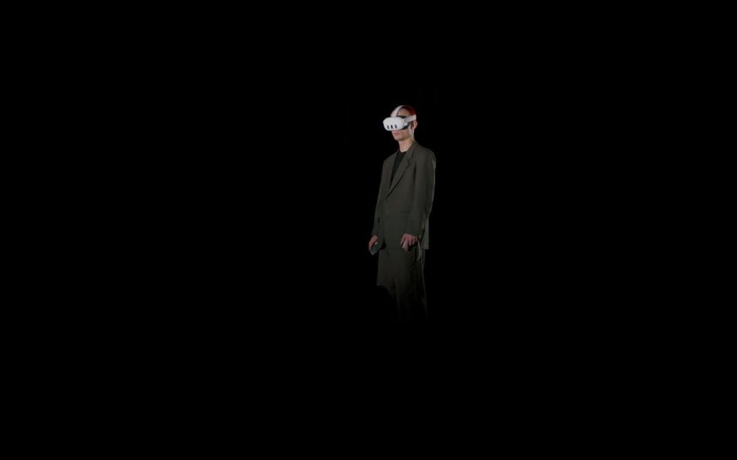 Film still from Alisa Berger’s film “RAPTURE”. Wearing a gray suit, a person stands in the darkness, a virtual reality headset on their head and a controller in each hand.