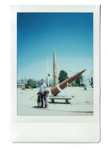 Polaroid of a man and two children in front of a missile display at an open-air museum.