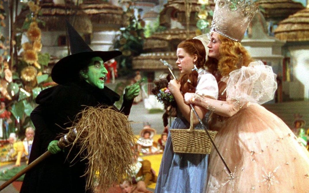 Filmstill aus THE WIZARD OF OZ: Ein Mädchen mit Zöpfen in einer Märchenszenerie: Hinter ihr ist eine Prinzessin mit Krone, vor ihr eine Hexe mit grün geschminktem Gesicht und schwarzem, spitzen Hut.