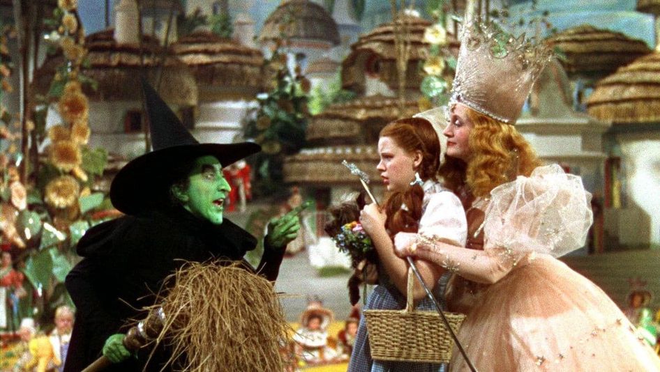 Film still from THE WIZARD OF OZ: A girl with pigtails in a fairy-tale setting: Behind her is a princess wearing a crown; in front of her is a witch with a face painted green and a black, pointed hat.