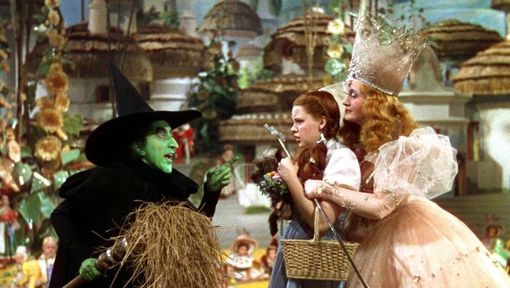 Film still from THE WIZARD OF OZ: A girl with pigtails in a fairy-tale setting: Behind her is a princess wearing a crown; in front of her is a witch with a face painted green and a black, pointed hat.