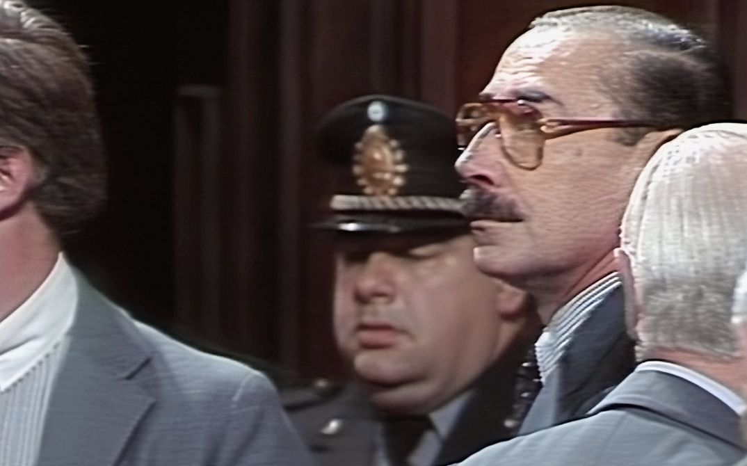 Still from the film "El juicio" by Ulises de la Orden. Several men in suits are standing next to each other, the one in the middle is wearing large glasses. There is another man in the background wearing a military uniform.