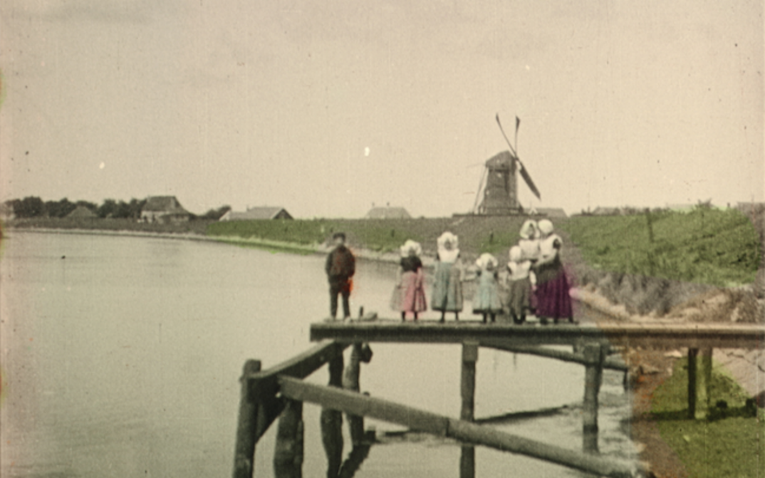 Filmstill aus DEAREST FIONA: Altes Bild einer Familie auf einem hölzernen Steg. In der Ferne sieht man eine Mühle und einige Häuser. Die Kleidung, die Vegetation und das Wasser sind handkoloriert. Der Rest ist schwarz-weiß.