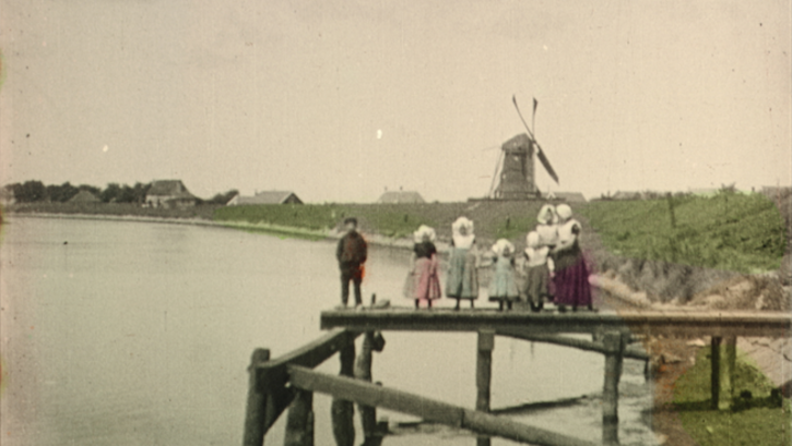 Film still from DEAREST FIONA: Old image of a family on a wooden pontoon. In the distance, we see a mill and some houses. The clothes, the vegetation and the water are hand colored. The rest is in black and white.