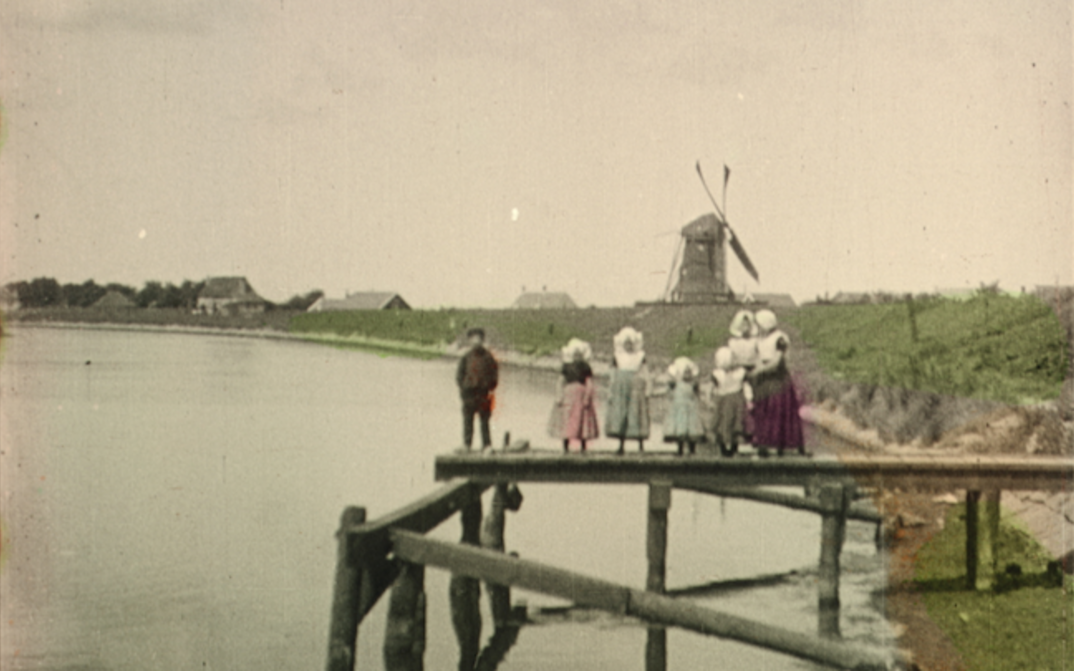 Filmstill aus DEAREST FIONA: Altes Bild einer Familie auf einem hölzernen Steg. In der Ferne sieht man eine Mühle und einige Häuser. Die Kleidung, die Vegetation und das Wasser sind handkoloriert. Der Rest ist schwarz-weiß.