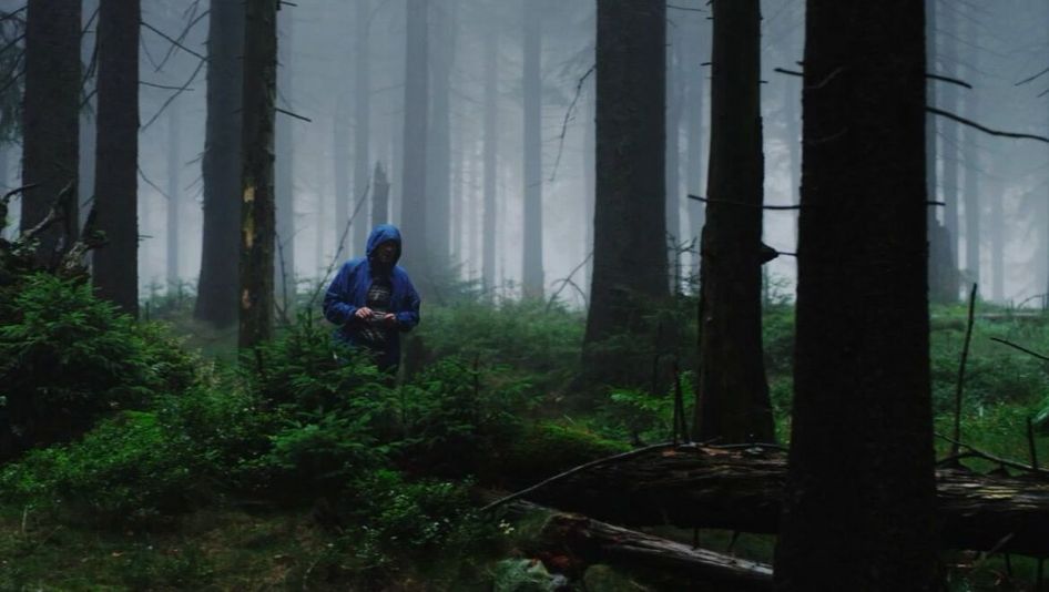 Filmstill aus EINE MINUTE DUNKEL: In einem nebligen Wald steht zwischen den Bäumen eine Person mit blauer Jacke und aufgesetzter Kapuze. Das Gesicht ist nicht zu erkennen.