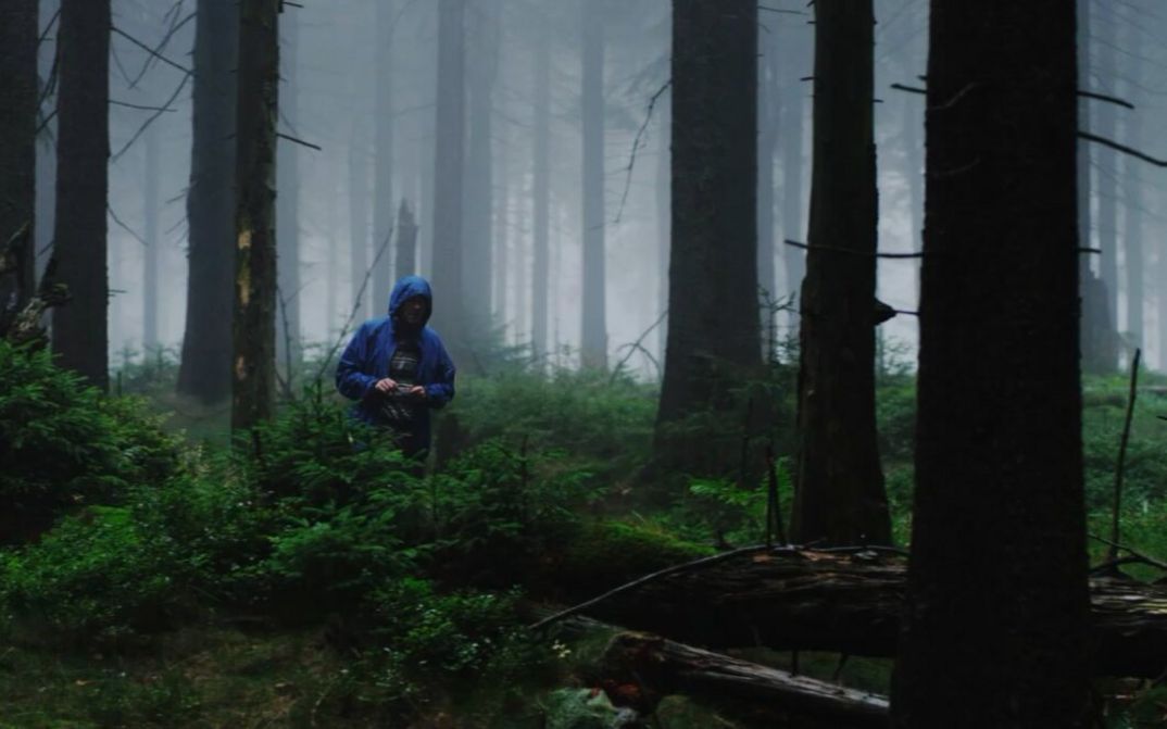Filmstill aus EINE MINUTE DUNKEL: In einem nebligen Wald steht zwischen den Bäumen eine Person mit blauer Jacke und aufgesetzter Kapuze. Das Gesicht ist nicht zu erkennen.