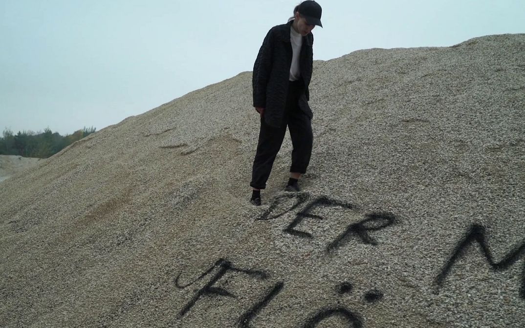 Filmstill aus ENDARCHIV: Eine Person steht auf einem Sandhügel, auf den etwas geschrieben wurde.