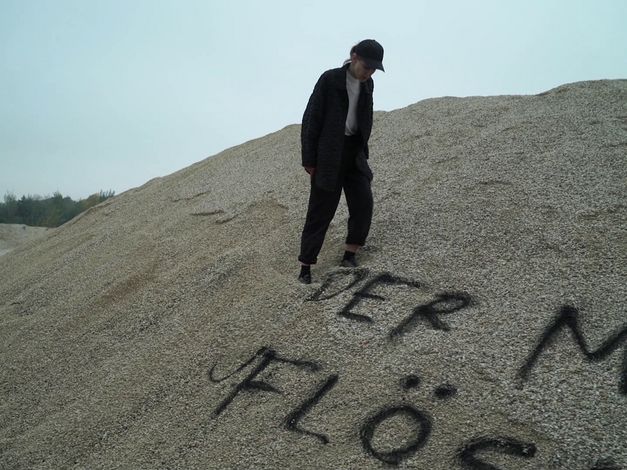 Filmstill aus ENDARCHIV: Eine Person steht auf einem Sandhügel, auf den etwas geschrieben wurde.