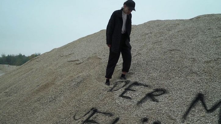 Filmstill aus ENDARCHIV: Eine Person steht auf einem Sandhügel, auf den etwas geschrieben wurde.