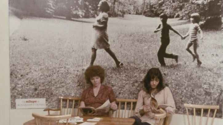 Film still from KAMPF UM EIN KIND. Two women sit at a table, on the wall behind them hangs a large photograph of a woman and two children in the grass.