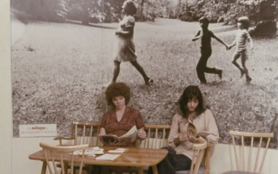 Film still from KAMPF UM EIN KIND. Two women sit at a table, on the wall behind them hangs a large photograph of a woman and two children in the grass.