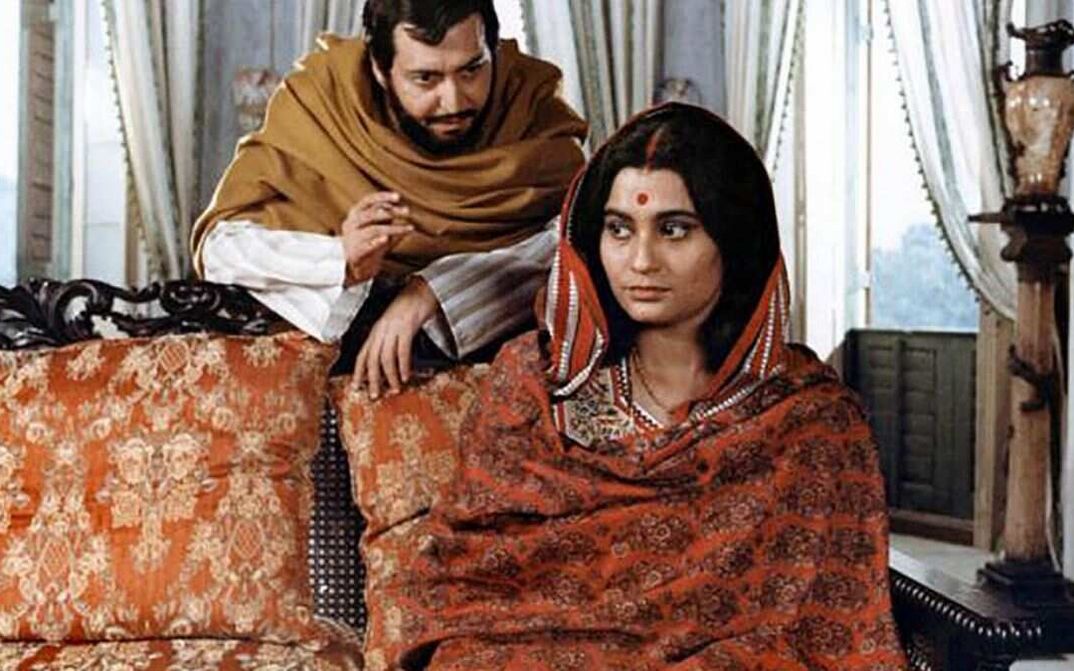 Filmstill aus GHARE BAIRE: Eine Frau sitzt auf einem Sofa, hinter ihr steht ein Mann und beugt sich zu ihr.