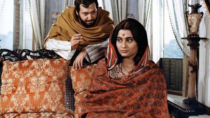 Film still from GHARE BAIRE: A woman sits on a sofa, a man stands behind her and leans towards her.