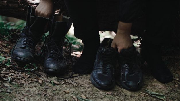 Filmstill aus dem Film „Black Strangers“ von Dan Guthrie. Rechts ein paar Schuhe, von einer Hand gehalten; links ein Paar Stiefel, von der anderen Hand gehalten.
