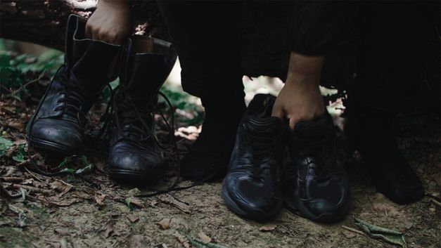 Film still from Dan Guthrie’s „Black Strangers“. To the right a pair of shoes, held by a hand; to the left a pair of Boots, held by the other Hand.