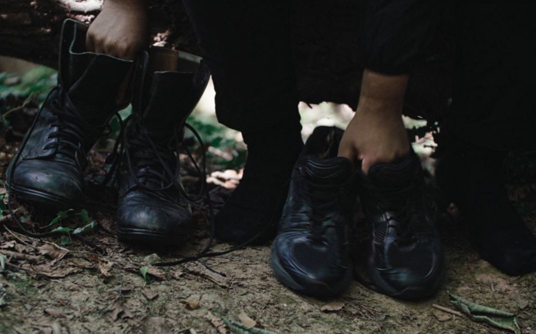 Film still from Dan Guthrie’s „Black Strangers“. To the right a pair of shoes, held by a hand; to the left a pair of Boots, held by the other Hand.