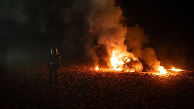 Film still from Riar Rizaldi’s film “Mirage: Eigenstate”. On a sandy expanse, a violent fire burns. In the foreground, a figure wearing a full-body suit and mask stares into the lens.