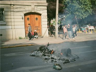 35-mm-Farbfoto. Junge Menschen stehen beziehungsweise gehen auf einem Bürgersteig, während vor ihnen die brennenden Überreste eines Fahrrads und andere nicht identifizierbare Objekte zu sehen sind.