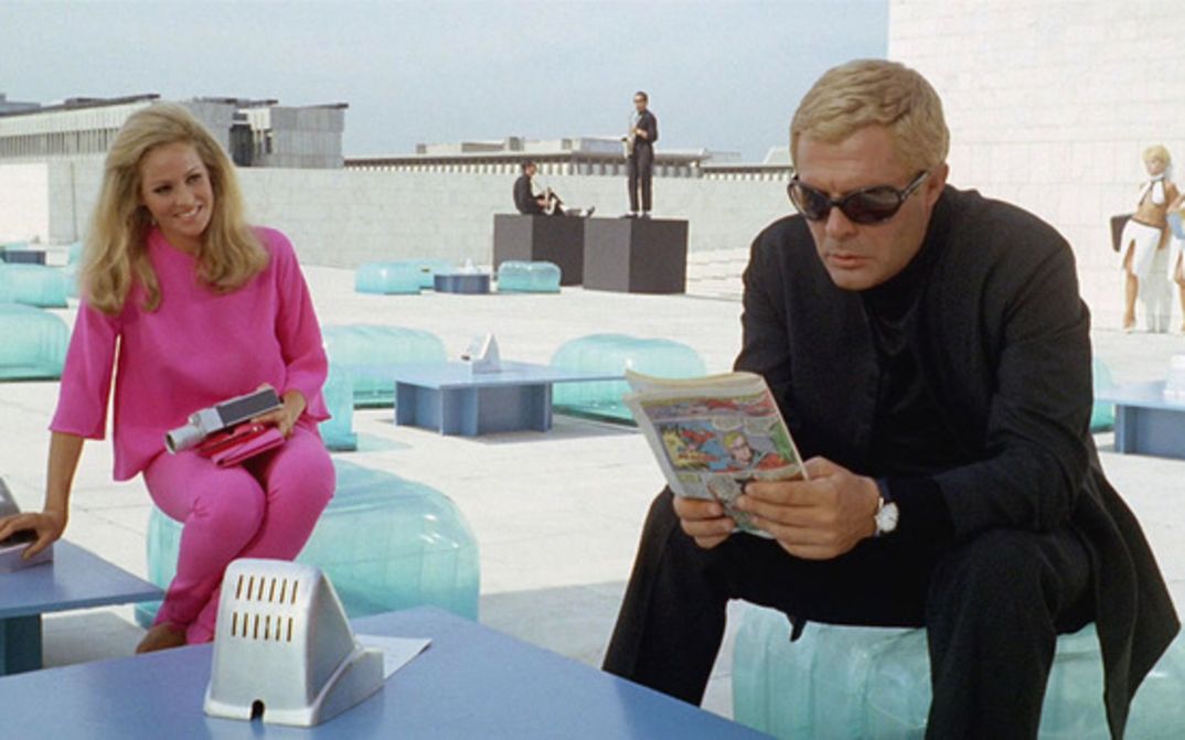 Film still from LA DECIMA VITTIMA: Marcello Mastroianni with blond hair and suit is sitting on the roof of a house in a futuristic setting. Next to him is a blonde woman dressed all in pink.