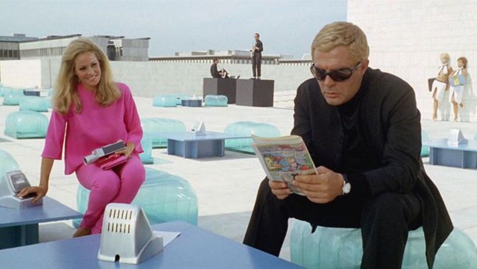 Filmstill aus LA DECIMA VITTIMA: Marcello Mastroianni mit blondiertem Haar und Anzug sitzt in einem futuristischen Ambiente auf dem Dach eines Hauses. Neben ihm ist eine blonde Frau, die ganz in Pink gekleidet ist.