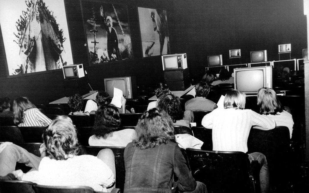 Takahiko Iimura „Time Tunnel: Takahiko Iimura at Kino Arsenal, 18. April 1973“. Ein Kinosaal in Schwarz-Weiß. Rückenansicht von Zuschauer*innen vor einer Reihe von Videomonitoren. An der linken Wand hängen drei große Poster. 