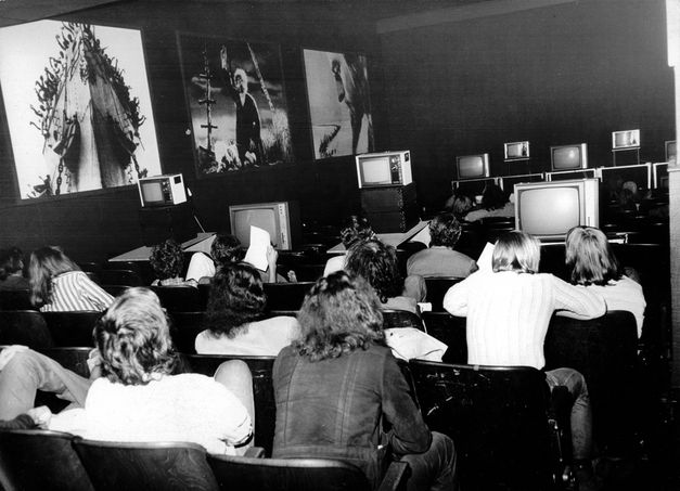 Takahiko Iimura „Time Tunnel: Takahiko Iimura at Kino Arsenal, 18. April 1973“. Ein Kinosaal in Schwarz-Weiß. Rückenansicht von Zuschauer*innen vor einer Reihe von Videomonitoren. An der linken Wand hängen drei große Poster. 