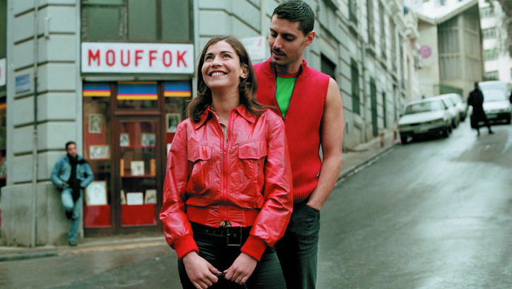 Film still from VIVA LALDJERIE: A young woman in a red leather jacket stands laughing on a busy street. A man stands behind her and looks at her.