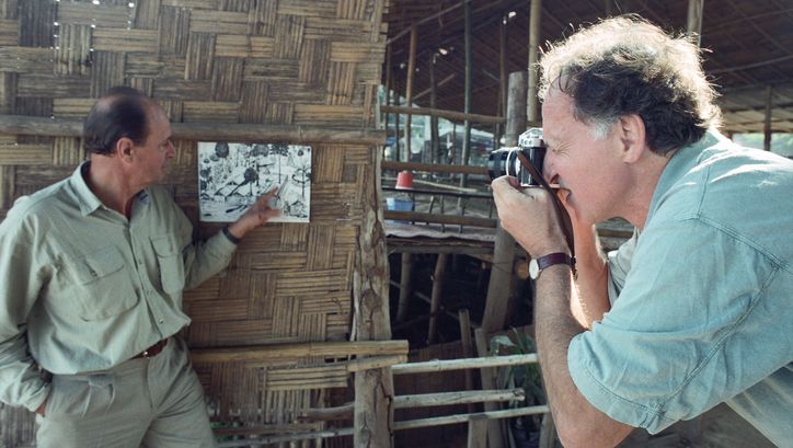 Behind the scenes of the film LITTLE DIETER NEEDS TO FLY: Werner Herzog is taking a photo of Dieter Denger who is pointing at a photograph pinned to a wooden wall.
