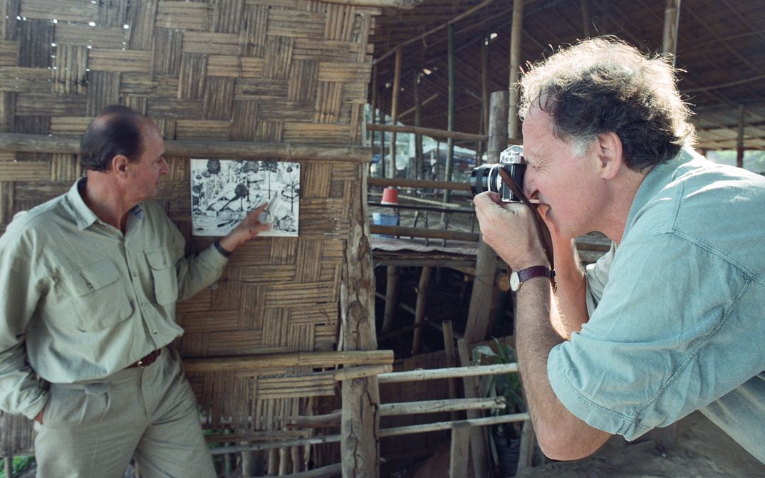 Behind the scenes of the film LITTLE DIETER NEEDS TO FLY: Werner Herzog is taking a photo of Dieter Denger who is pointing at a photograph pinned to a wooden wall.