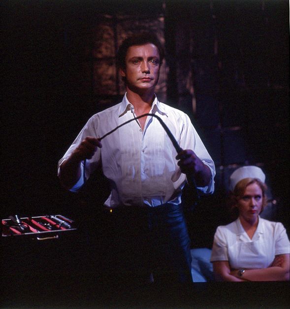 Mann in weißem Hemd hält ein Peitschenähnliches Objekt, Frau in Krankschwestertracht sitzt im Hintergrund.