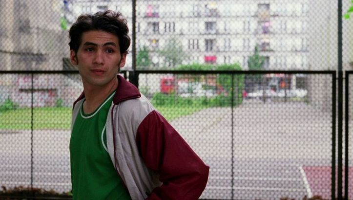 Film still from DEALER: A man in a tracksuit jacket stands in front of a fence and looks to the right. Cars, a building and green spaces can be seen in the background.