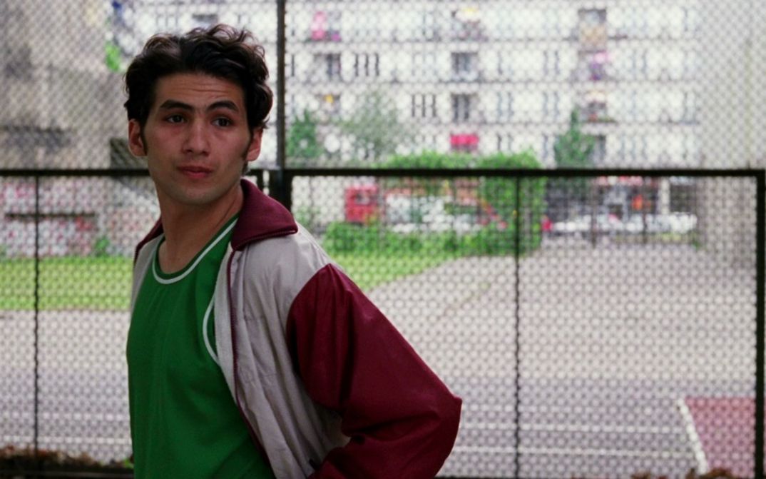 Filmstill aus DEALER: Ein Mann in Trainingsjacke steht vor einem Zaun und schaut nach rechts. Im Hintergrund sind Autos, ein Gebäude und Grünflächen zu erkennen.