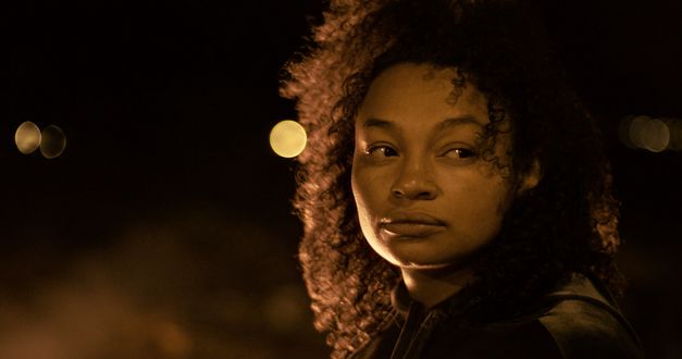 Still from the film "Mato seco em chamas (Dry Ground Burning)" by Adirley Queirós and Joana Pimenta. A close-up of a woman at night. Some lights in the background. 