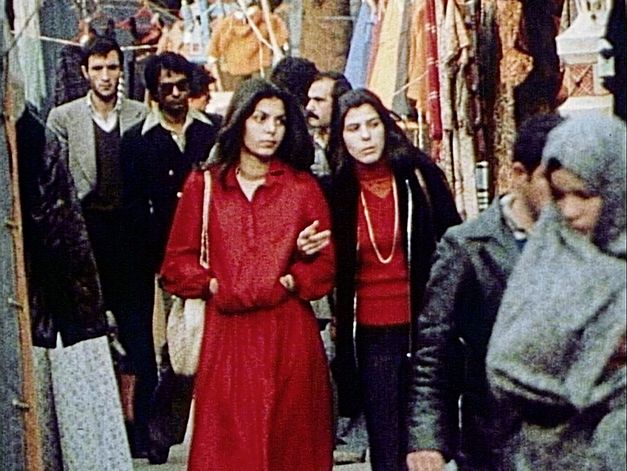 Still from the film "Între revoluții" by Vlad Petri. Two women, one dressed entirely in red, the other with a red shirt and a dark jacket, are walking along a market together.