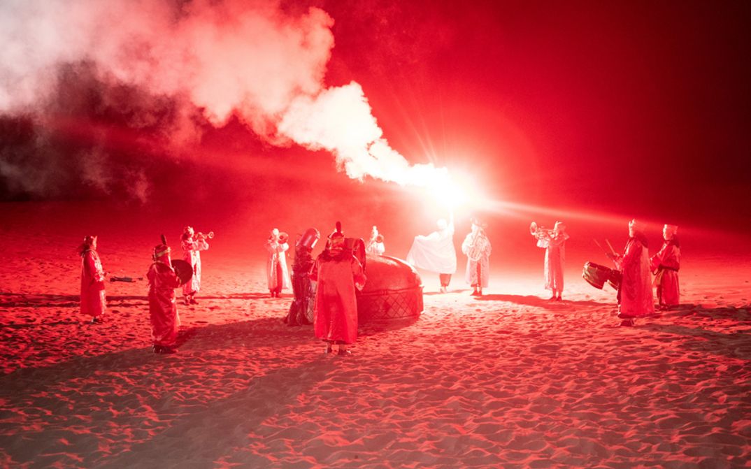 Film still from Riar Rizaldi’s film “Mirage: Eigenstate”. On a sandy, desert plateau bathed in red light, a mystical ceremony unfolds. Silhouettes form a circle, musical instruments in hand. At the center, a solitary figure rises, raising towards the sky an incandescent staff that releases swirls of smoke into the night.