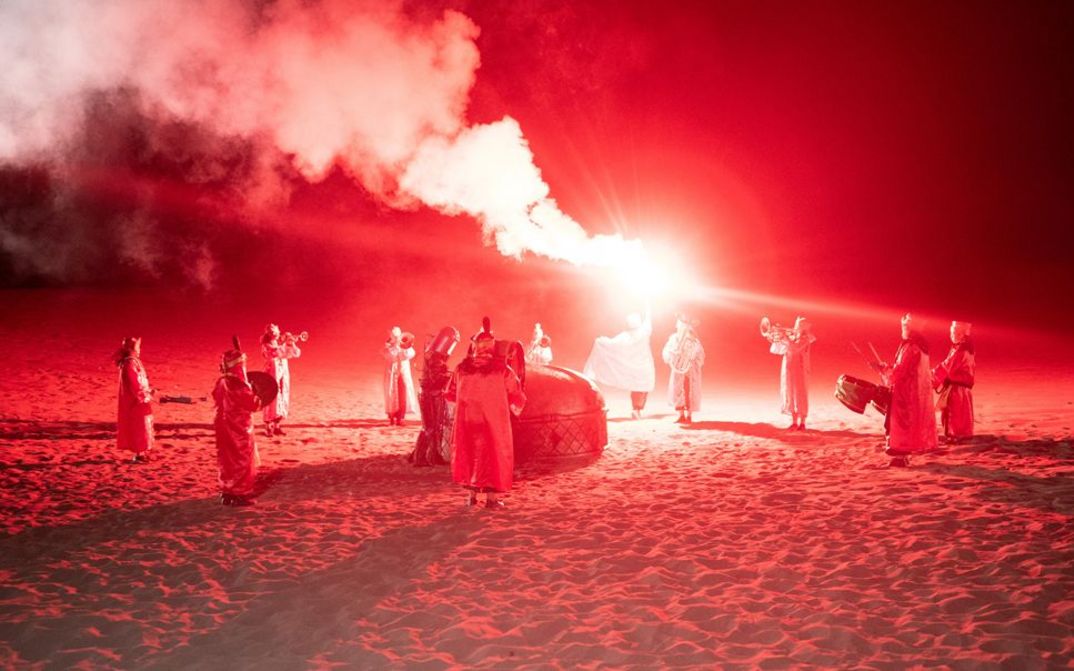 Film still from Riar Rizaldi’s film “Mirage: Eigenstate”. On a sandy, desert plateau bathed in red light, a mystical ceremony unfolds. Silhouettes form a circle, musical instruments in hand. At the center, a solitary figure rises, raising towards the sky an incandescent staff that releases swirls of smoke into the night.