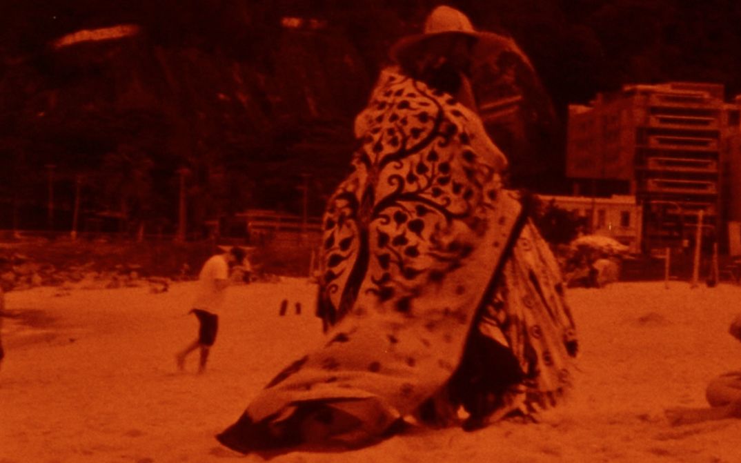 Film still from TO BRASIL: Monochrome red colored picture of a person with an eye-catching cape on the beach.