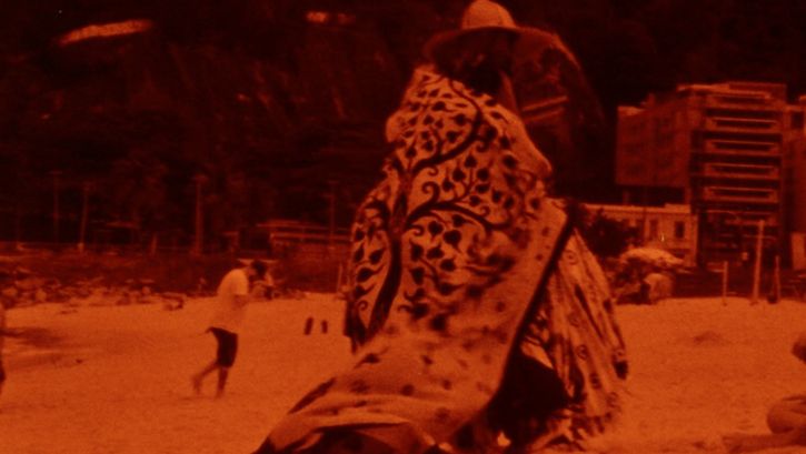Film still from TO BRASIL: Monochrome red colored picture of a person with an eye-catching cape on the beach.