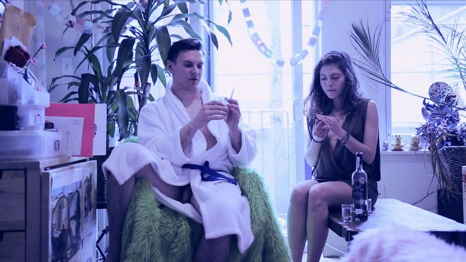 Filmstill aus OBJECT: Ein junger Mann und eine junge Frau sitzen in einem Zimmer. Er trägt nur einen weißen Bademantel, der halb offen ist. Vor ihr sind eine Flasche Alkohol und zwei Gläser.