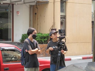 A behind-the-scenes shot of two people. One is filming and the other is standing next to them, wearing a face mask. There is a red car behind them.