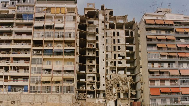 Film still from NOS GUERRES IMPRUDENTES. View of three apartment blocks, the middle one destroyed.