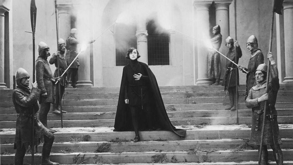 Filmstill aus HAMLET: Asta Nielsen als Hamlet steht ganz in Schwarz gekleidet und mit einem langen Umhang auf der Treppe eines prächtigen Hauses. Links und rechts auf der Treppe stehen aufgereiht Wachmänner.